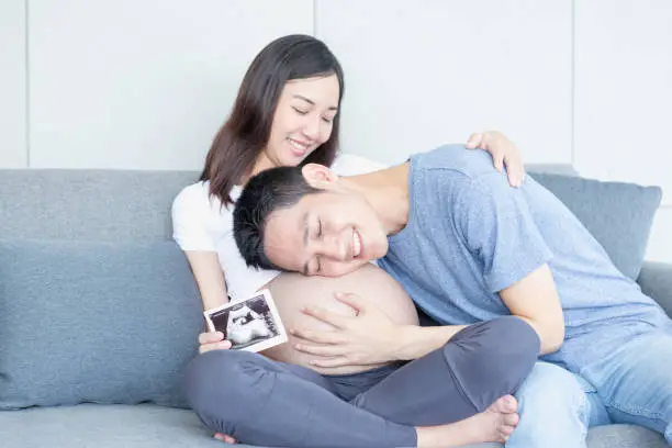 Photo of Father is kissing his mother's stomach. Young husband kissing his pregnant wife's tummy in living room with the baby ultrasound photo.