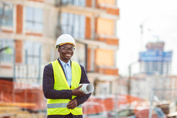 Portrait of African American man architect wearing a vest and helmet Portrait of African American man architect wearing a vest and helmet, he stands and holding a blueprints with the sky and urban in background. Engineer and architect concept. project manager stock pictures, royalty-free photos & images