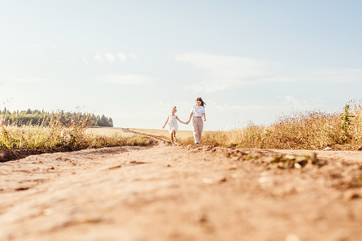 Beautiful young mother and her daughter having fun and walk at the country road. Ground on foreground, sky background. White clothes, freedom concept.