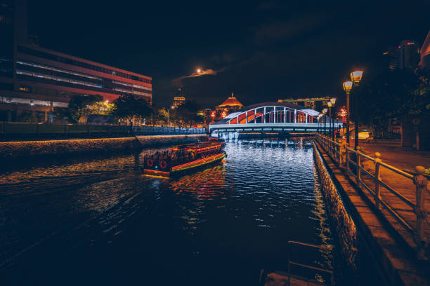Singapore River and Boat Quay area of Singapore seen at night. In the far back the Marina Bay Sands hotel is showing behind a bridge. A boat passing by on the river.