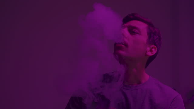 Portrait of a young man, blowing the stack of smoke in the room, illuminated with purple light