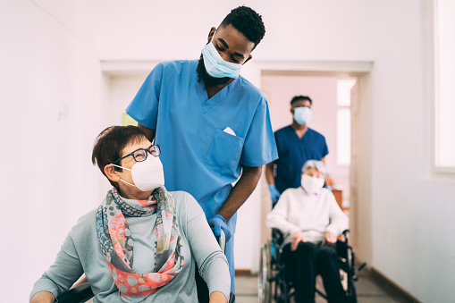 Dos pacientes femeninas de alto rango en sillas de ruedas, empujados por trabajadores sanitarios masculinos a lo largo de un pasillo, todos llevan máscaras faciales protectoras photo