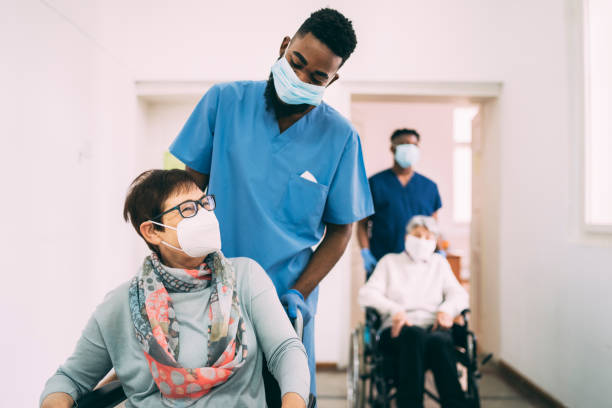 two senior female patients in wheelchairs, being pushed by male healthcare workers along a corridor, all are wearing protective face masks - male nurse black nurse doctor imagens e fotografias de stock