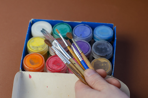A man's hand holding several paint brushes. A set of paints in a box in the background. Homemade leisure, art, painting concept.