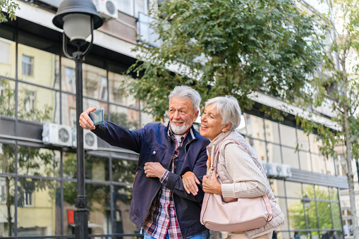An Older Man and his Happy Wife are Feeling Enjoyment During a Walk in the Streets of Large City and Photographing with a Mobile Phone.