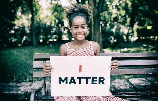 African american girl sitting on the bench outdoors and looking at the camera. She's showing an message that says I matter.