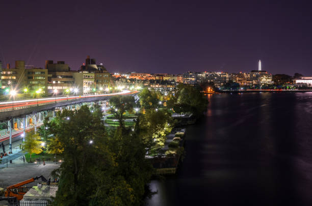 Night view of an illuminated waterfront park under an elevated road with high rise residential and office buildings in background. Washington DC.