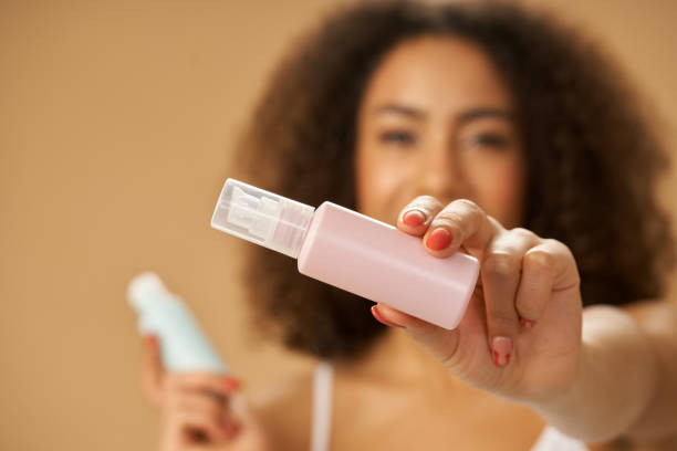 primer plano de la joven de raza mixta que sostiene una botella rosa con producto de belleza mientras posa aislada sobre el fondo beige. concéntrate en una botella - frizzy 20s adult african descent fotografías e imágenes de stock