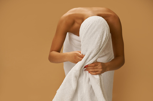 Young woman after shower wipes her hair with a white towel while standing isolated over beige background. Beauty, spa treatment concept