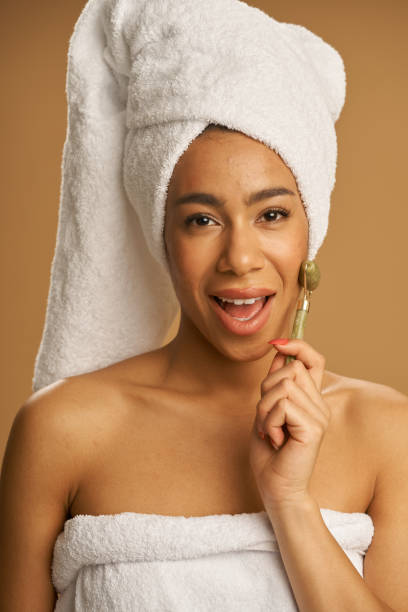 istock Playful mixed race young woman after shower looking at camera while using jade roller for massaging her face isolated over beige background 1312958389