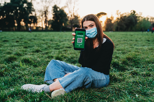 Young woman showing a Covid-19 vaccine passport on her smart phone to the camera. She's outdoor in a public park. She's wearing a protective face mask. Dummy QR code on the screen.
