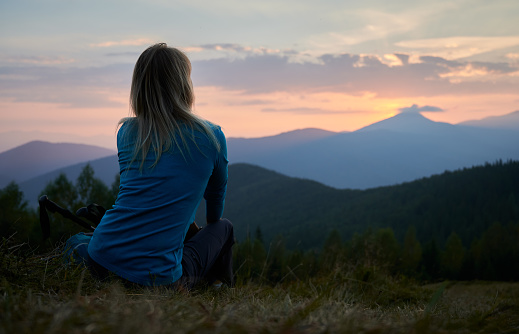 Back view of female hiker who resting after day of hike and watching at sunset above mountain beskids. Tourist enjoying landscape of mountain hills and evening sky when sun setting over horizon.