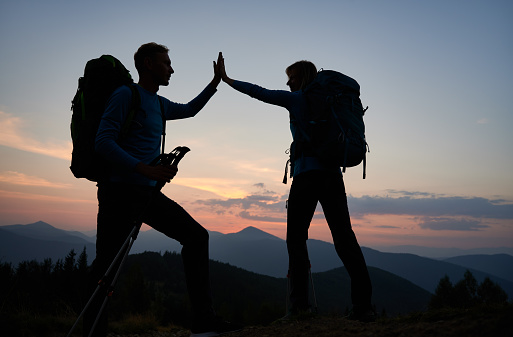 Full length of young man and woman travelers giving high five while hiking together in the mountains during sunset. Concept of travelling, hiking and relationships.