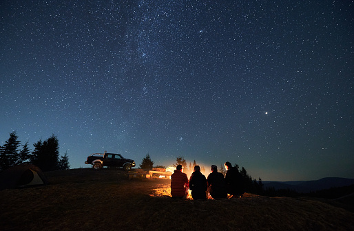 Evening starry sky over mountain valley with car and hikers near campfire. Group of travelers sitting near bonfire under majestic blue sky with stars. Concept of night camping, hiking and travelling.