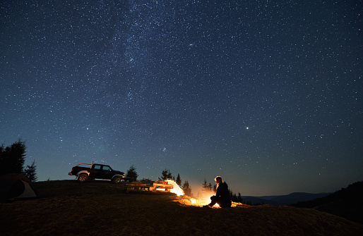 Female camper resting by bonfire and enjoying pleasant atmosphere in camp outdoor. Two tents, wooden table between them and black jeep parked on hill. Beautiful shining stars in the night sky.