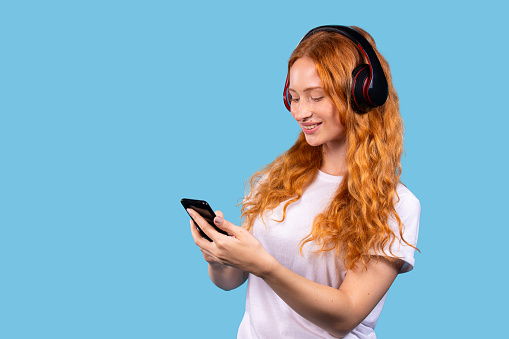 Woman listening to music via wireless headphones using mobile smartphone, chatting in social networks, posing on blue background with empty space for info. Life, time to relax. . High quality photo
