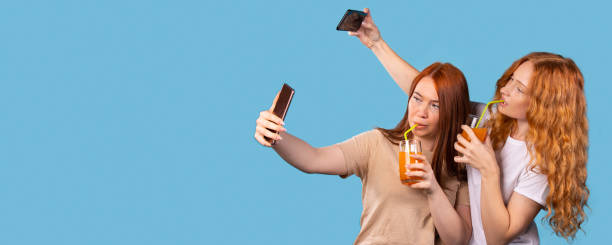 banner, formato longo no azul. imagem de jovens felizes e emocionais duas amigas bebendo coquetéis sentados dentro de casa vestidas com camisetas de verão tiram uma selfie pela câmera frontal do smartphone. anúncio de texto espacial - outdoors drinking women friendship - fotografias e filmes do acervo