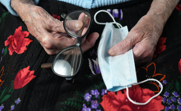 a 90-year-old grandmother holds a protective mask against the covid-19 coronavirus and eyeglasses. view from above. protecting older people in quarantine. - 80 year old imagens e fotografias de stock