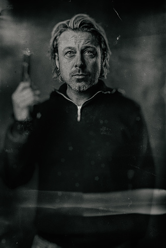 Vintage wet plate portrait of blond man with stubble beard and handgun in sweater.