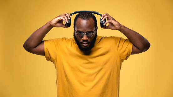 African-american ethnicity person with eyeglasses and wireless black headphones is listening music. Concept for dj, techno, pop, rap, r&b music photography. Studio portrait on yellow background
