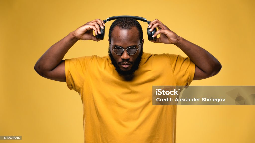 African-american ethnicity person with eyeglasses and wireless black headphones is listening music. Concept for dj, techno, pop, rap, r&b music photography. Studio portrait on yellow background DJ Stock Photo