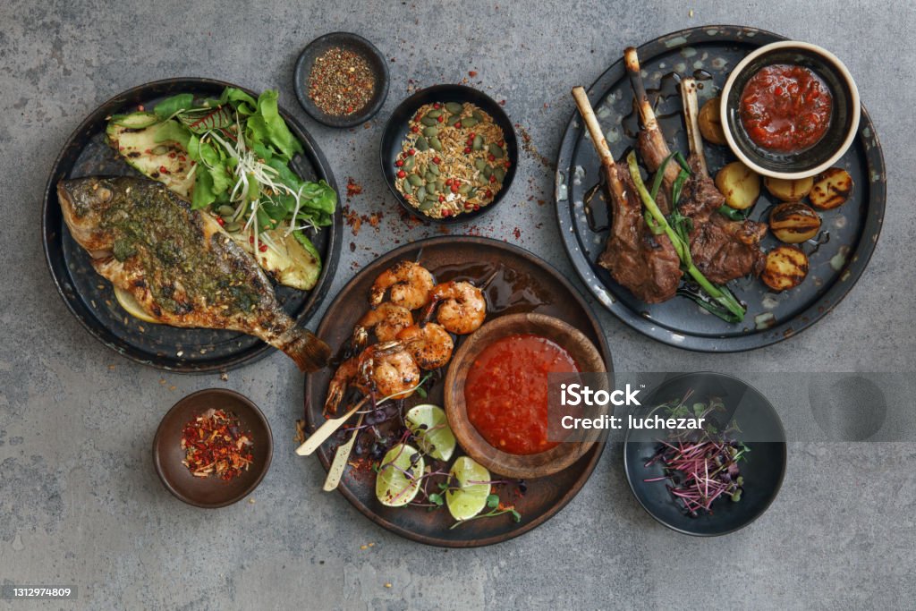 Latino Food Brazilian coconut shrimp kebabs. Barbecued sea bream with grilled vegetables. Brazilian grilled lamb chops. Flat lay top-down composition on concrete background. Barbecue Grill Stock Photo