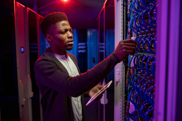 Black man examining datacenter equipment Concentrated young Black man using tablet while examining datacenter equipment in dark server room man and machine stock pictures, royalty-free photos & images