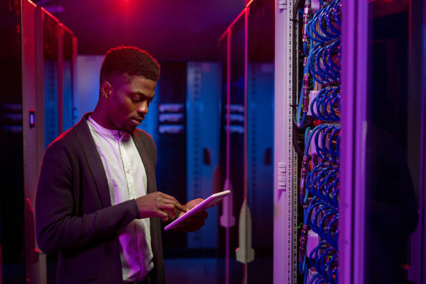 Setting up system at data center Serious young African-American data engineer standing at open server rack cabinet and using tablet while setting up system at data center mainframe stock pictures, royalty-free photos & images