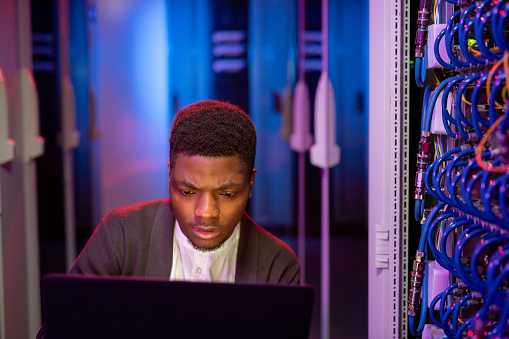 Busy young black IT engineer using modern laptop in server room while administrating computer network