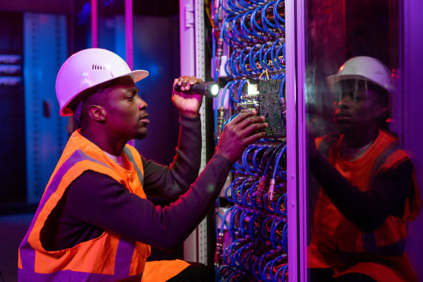 Black workman fixing server problem Concentrated Black workman in hardhat and orange vest crouching by server and using flashlight while fixing server problem electrician stock pictures, royalty-free photos & images