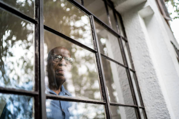 uomo anziano che guarda attraverso la finestra di casa - solitario foto e immagini stock