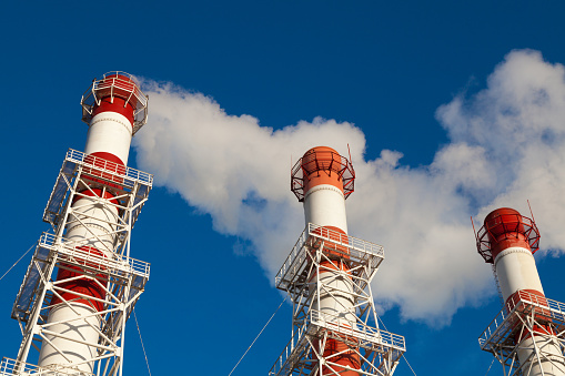 Three striped red and white factory chimneys from which white smoke comes out, stand against a blue sky