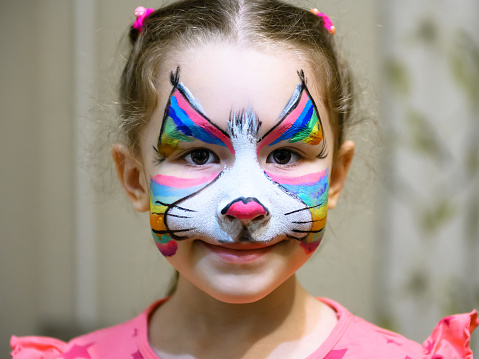 Kid with face painting of kitty, cute little girl with painted mask on her face of rainbow cat. Portrait of pretty child with beautiful makeup, unique facial body art for party. Colorful draw on skin.