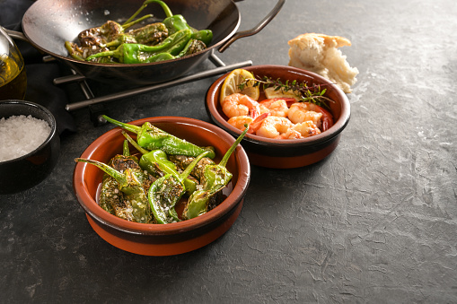 Spanish tapas, fried pimientos or padron peppers and shrimps in traditional bowls on a dark gray slate background, copy space, selected focus, narrow depth of field