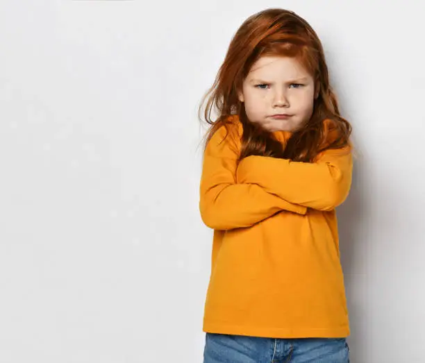 Serious small red haired girl in yellow comfortable longsleeve standing with crossed hands and frowning looking at camera over white background, copy space. Happy childhood, stylish look concept