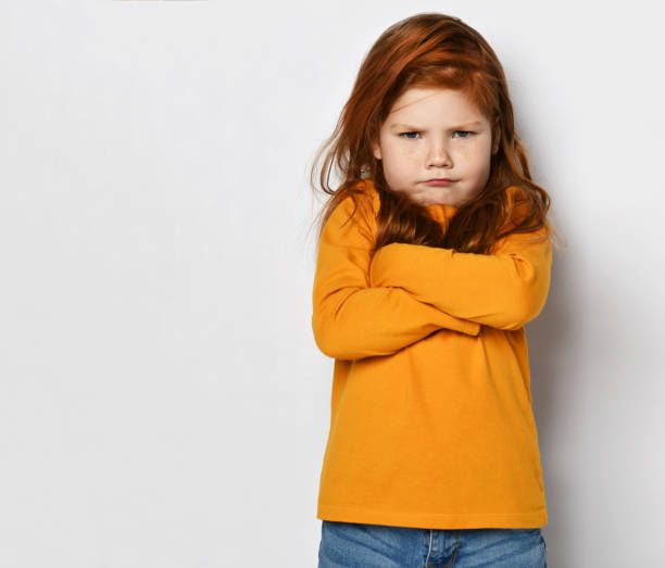 serious small red haired girl in yellow comfortable longsleeve standing with crossed hands and frowning - frowning imagens e fotografias de stock