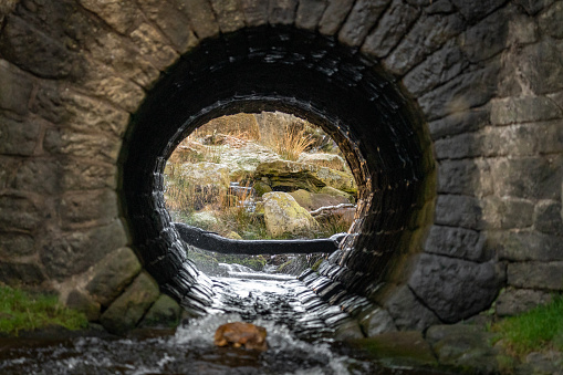 Old circular sewer brick culvert tunnel water river running through flowing, granite stone in Peak District countryside passing under road.