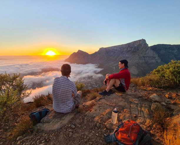 hikers at sunrise on table mountain Two men relaxing on top of a mountain at sunrise overlooking clouds, cape town city center and with Table Mountain as a backdrop table mountain south africa stock pictures, royalty-free photos & images