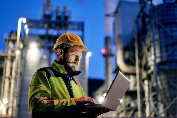 Male engineer using laptop during night shift. Young engineer wearing a helmet and using a laptop during his night shift. Engineering concept. power station stock pictures, royalty-free photos & images