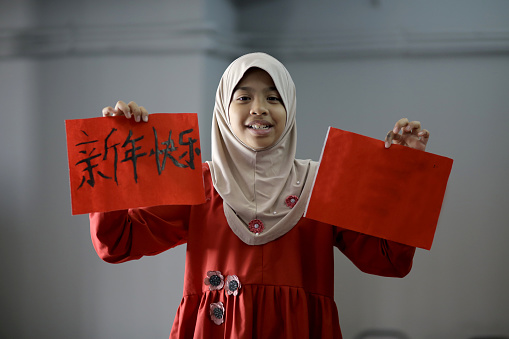 A Muslim girl is holding calligraphy written 'Happy Chinese New Year' and name 'Sufiya' in Chinese characters. Sufiya is a girl's name in Malay language.