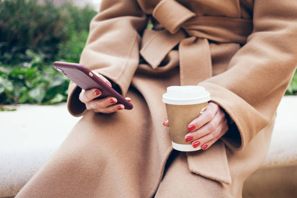 istock Close up of woman hands holding mobile phone, using internet while sitting outdoors on the bench and drinking coffee. 1313077352