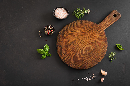 Empty wooden round board with herbs and spices on black stone kitchen table, top view, flat lay. Wooden pizza platter, copy space. Food background.