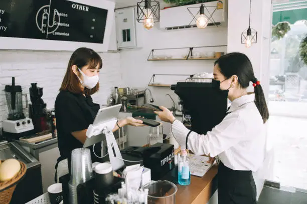 Photo of Asian woman cafe owner businesswoman receiving payment from her customer at counter using QR code contactless payment - stock photo