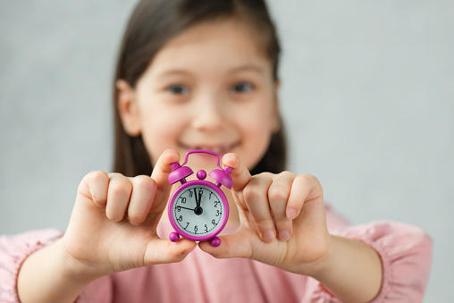 Little girl is smiling at camera with pink alarm clock in her hands. Clocks time is 5 to 12.