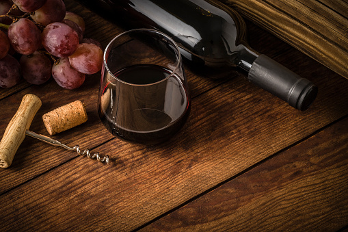 High angle view of a drinking glass full of red wine, a wine bottle, a cork, a screw cap and a branch of red grapes laying at the top left corner leaving a useful copy space at the lower right corner on a old fashioned wooden table. Predominant color is brown. Low key DSLR photo taken with Canon EOS 6D Mark II and Canon EF 100 mm f/ 2.8