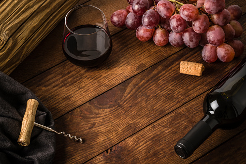 High angle view of a red wine bottle, a branch of grapes, a drinking glass full of wine and a screw cap disposed on a frame shape leaving a useful copy space at the center of the image on a old fashioned rustic wooden table. Predominant color is brown. Low key DSLR photo taken with Canon EOS 6D Mark II and Canon EF 100 mm f/ 2.8