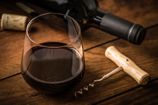 High angle view of a drinking glass full of red wine beside a wooden handle screw cap, a cork and a red wine bottle on a rustic wooden table. Predominant color is brown. Low key DSLR photo taken with Canon EOS 6D Mark II and Canon EF 100 mm f/ 2.8