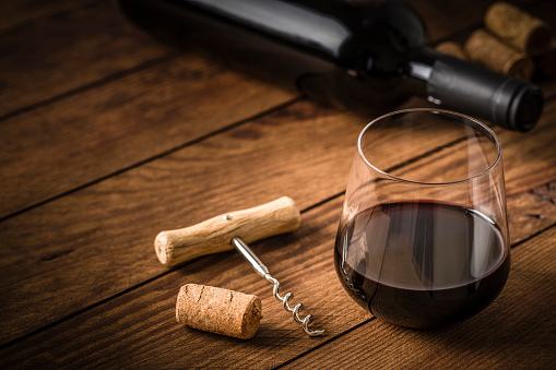 High angle view of a drinking glass full of red wine surrounded by a screw cap, a cork and red wine bottle. All the objects are at the right side of the image leaving a useful copy space at the left side on a rustic wooden table. Predominant color is brown. Low key DSLR photo taken with Canon EOS 6D Mark II and Canon EF 100 mm f/ 2.8