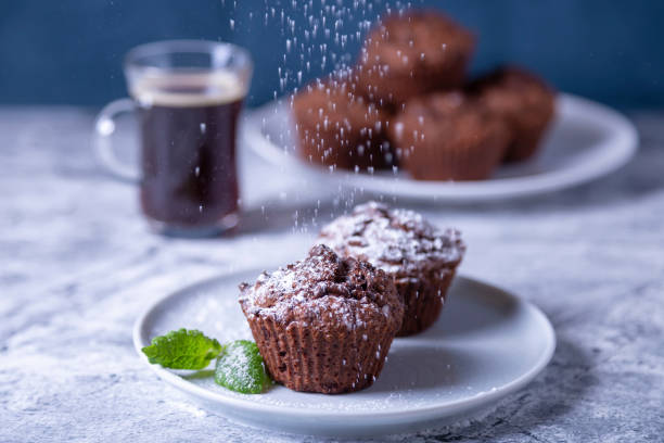 Chocolate muffins with mint on a white plate, sprinkled with powdered sugar. Homemade pastries. In the background is a cup of coffee and a plate of cupcakes. Chocolate muffins with mint on a white plate, sprinkled with powdered sugar. Homemade cakes. In the background is a cup of coffee and a plate of muffins. Marble table and blue background. Close-up. sprinkling powdered sugar stock pictures, royalty-free photos & images