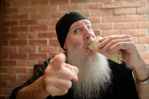 Portrait of cool mature man with long gray beard sitting indoors against brick wall background